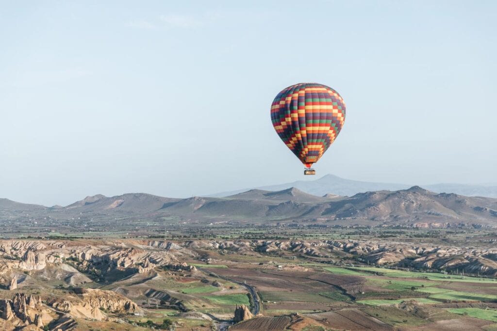 A single multicolored hot air balloon soars above a wide, rocky valley with winding paths and hills in the distance under a clear sky.