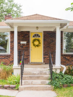 Charming brick fixer-upper with a bright yellow front door, front porch steps, and overgrown landscaping ready for renovation.