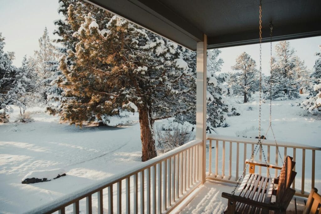 A peaceful snowy landscape viewed from a wooden porch with a swing bench, surrounded by trees blanketed in fresh snow.