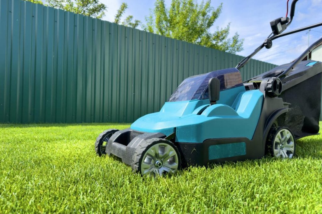 Battery-powered blue electric lawn mower on a green lawn, with a metal fence and trees in the background on a sunny day.