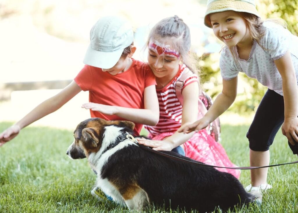Three young children gently pet a leashed dog on a grassy lawn, smiling and interacting carefully with the animal.