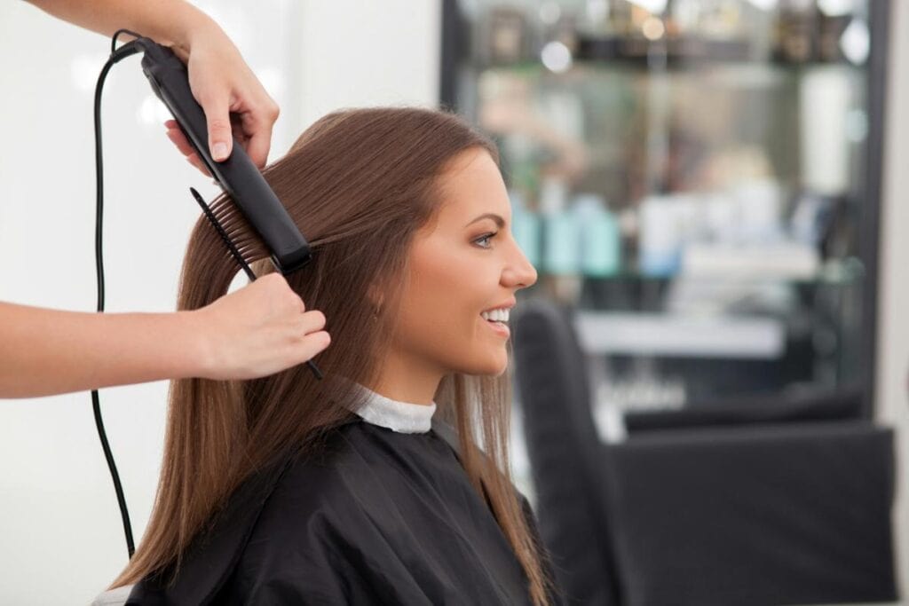 A stylist uses a flat iron and comb to straighten a woman’s long brown hair in a salon, while she smiles during the process.
