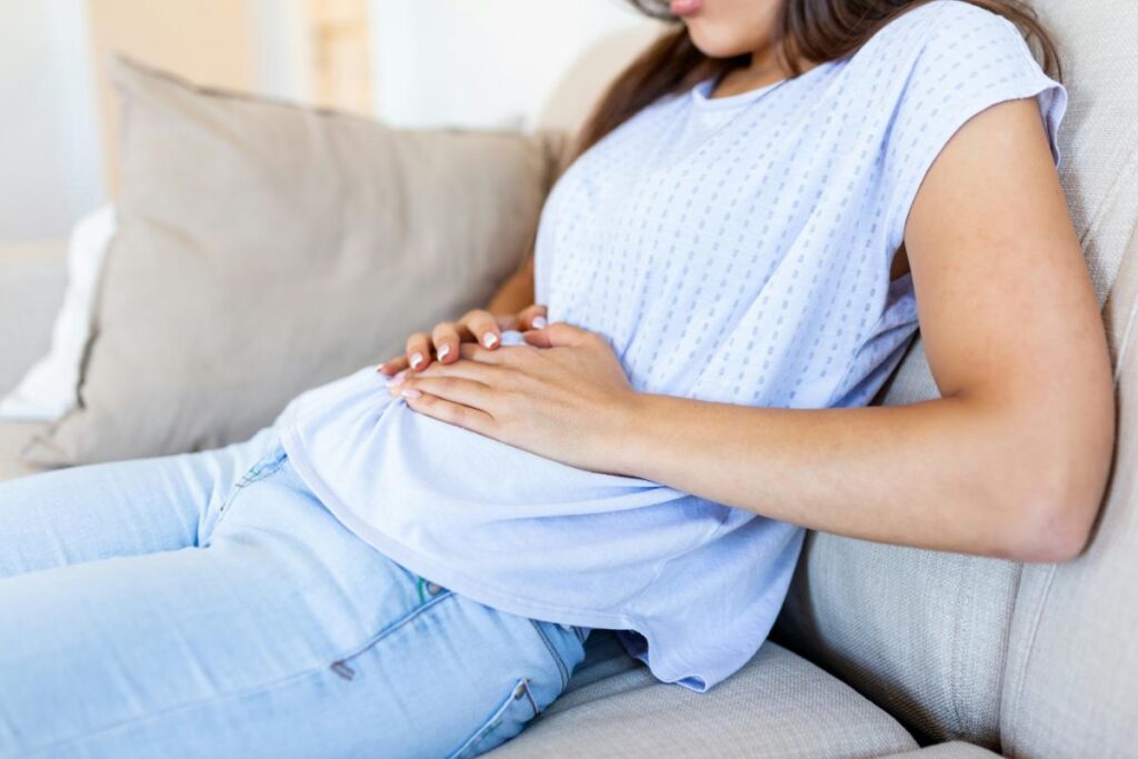 Woman sitting on a couch with hands on her abdomen, possibly experiencing bloating or digestive discomfort related to gut health.