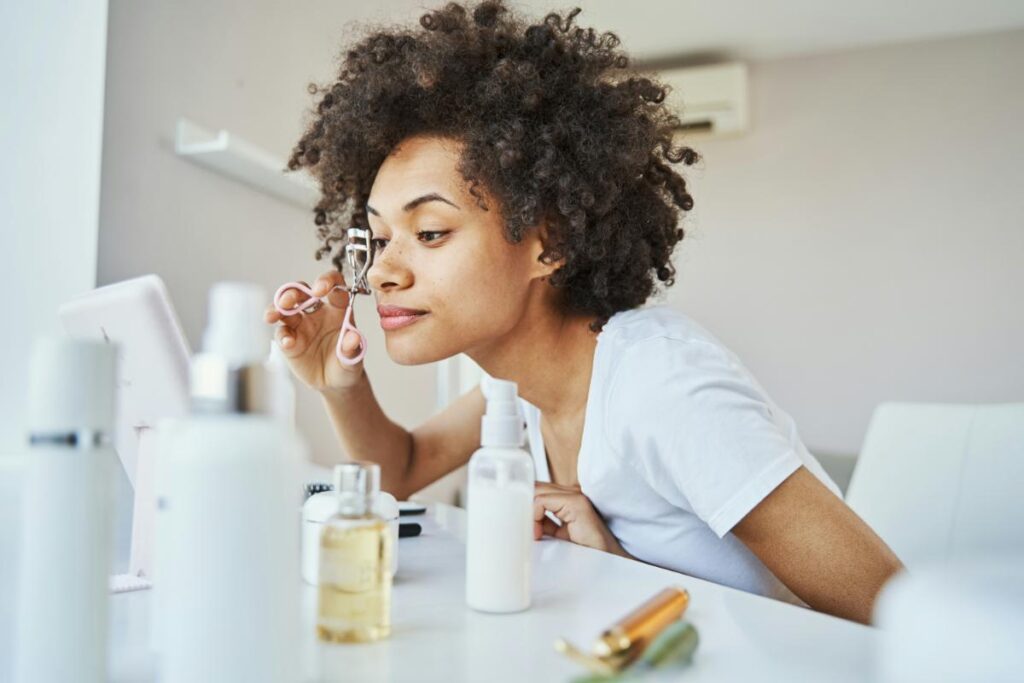 Young woman using an eyelash curler at home in front of a mirror, preparing her lashes as part of her beauty routine.