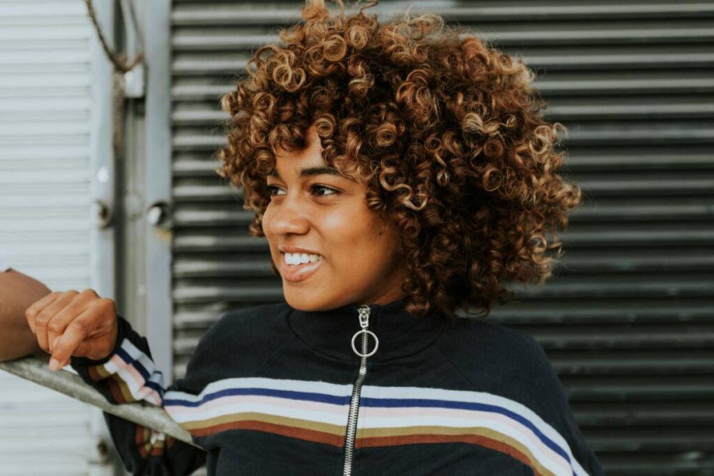 A woman with voluminous, defined curls smiles while standing outdoors, wearing a striped zip-up top.