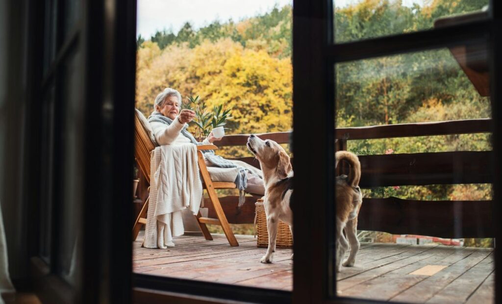 An elderly woman relaxes with a cup of tea on a cozy deck while her beagle looks up at her, enjoying a peaceful pet-friendly outdoor moment.