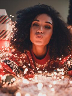 Woman wearing a festive sweater surrounded by glowing holiday lights and wrapped presents, blowing a kiss in a cozy, decorated room.