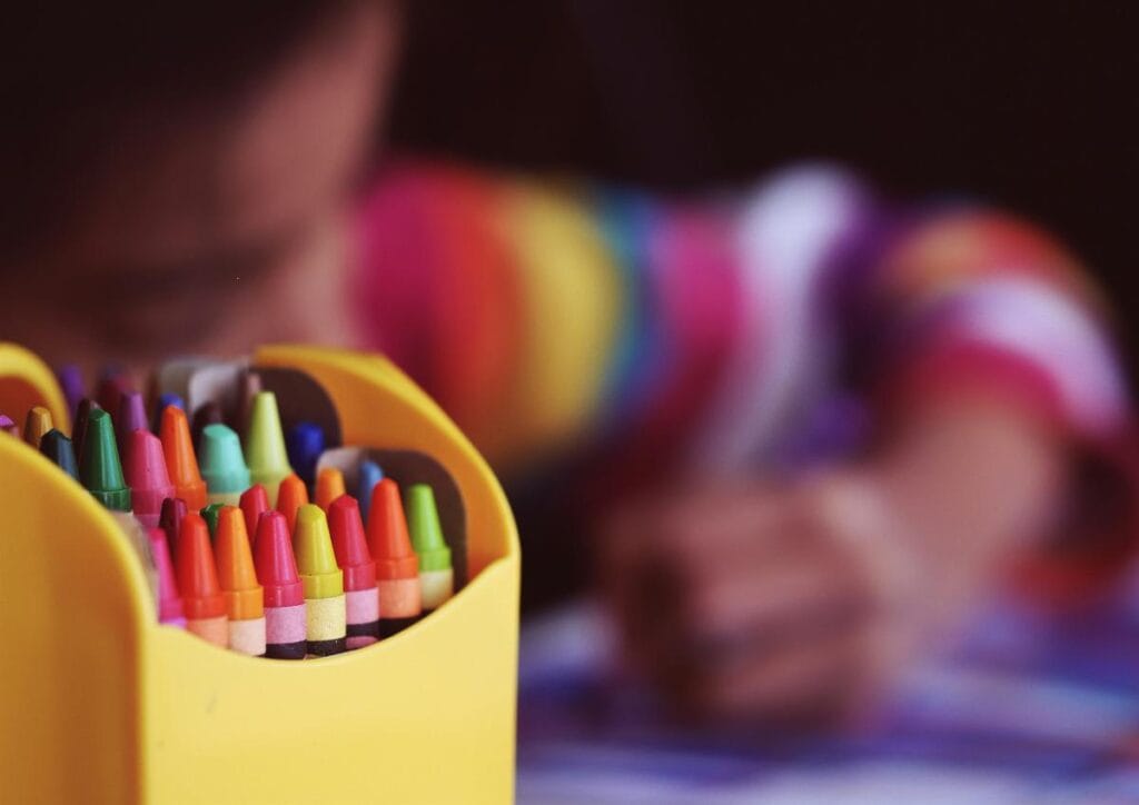 child coloring in blurred background with selective focus on box of crayons in foreground