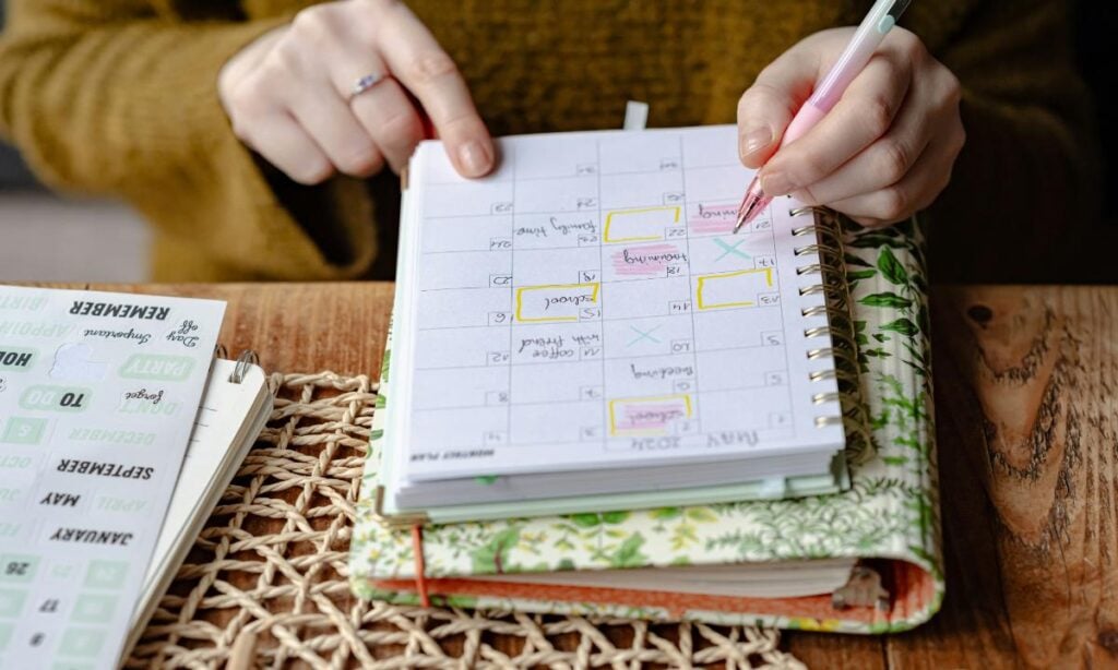 Close-up of a woman using a pen to organize a family schedule in a planner, surrounded by notebooks and stickers on a wooden table.