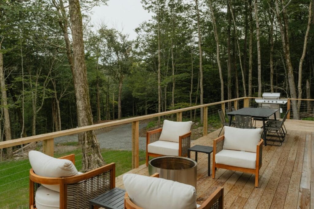 A spacious wooden deck with cushioned chairs, a grill, and forest views offers a stylish and functional outdoor space for relaxing and entertaining.