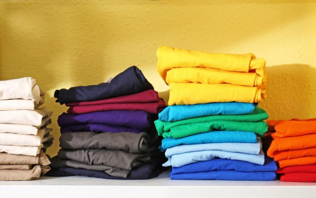 Stacks of neatly folded t-shirts in a variety of colors, arranged on a white shelf against a yellow wall.