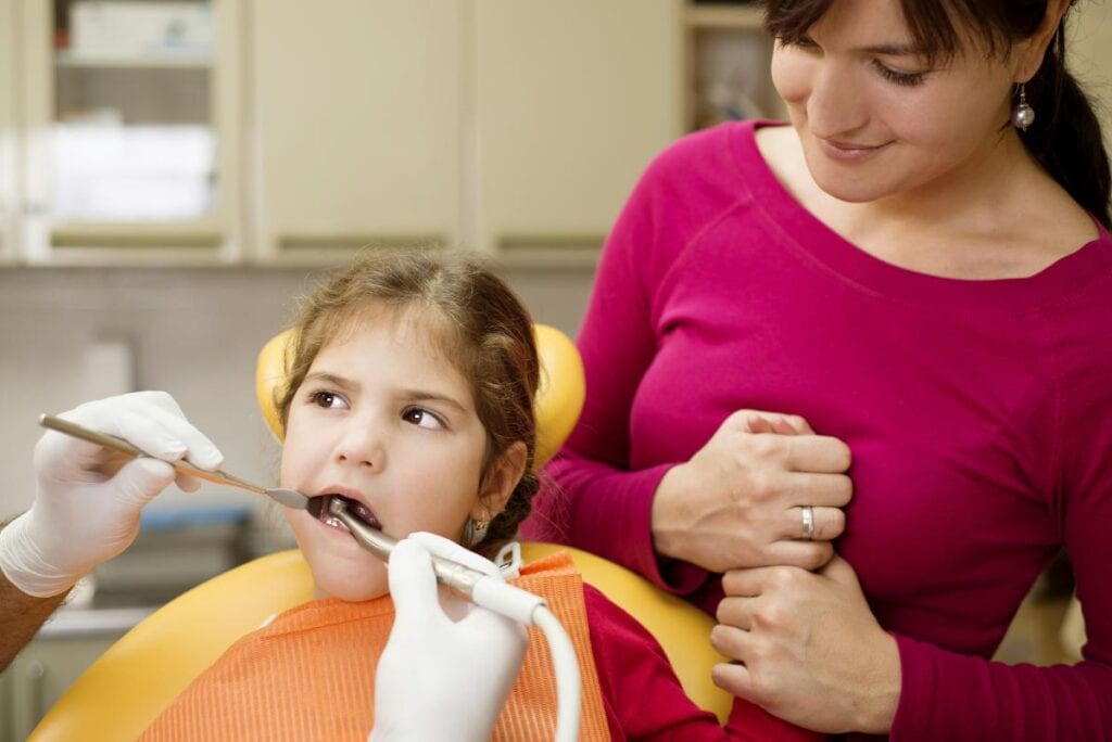 A young girl receives a dental checkup while her mother stands beside her supportively, showing the comfort of shared dental experiences.