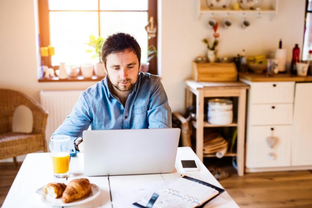 Man working on a laptop at a bright kitchen table, surrounded by natural light&mdash;highlighting how light and space impact mood and productivity at home.