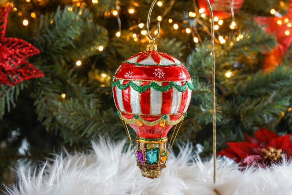 Festive red and white striped hot air balloon ornament with glittery details hanging on a Christmas tree with twinkling lights.