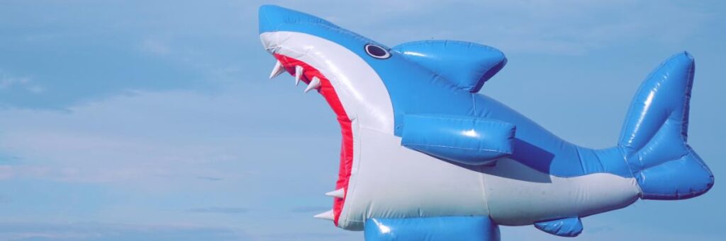 A large inflatable shark with an open red mouth and white teeth stands tall under a partly cloudy sky, creating a bold and eye-catching display.