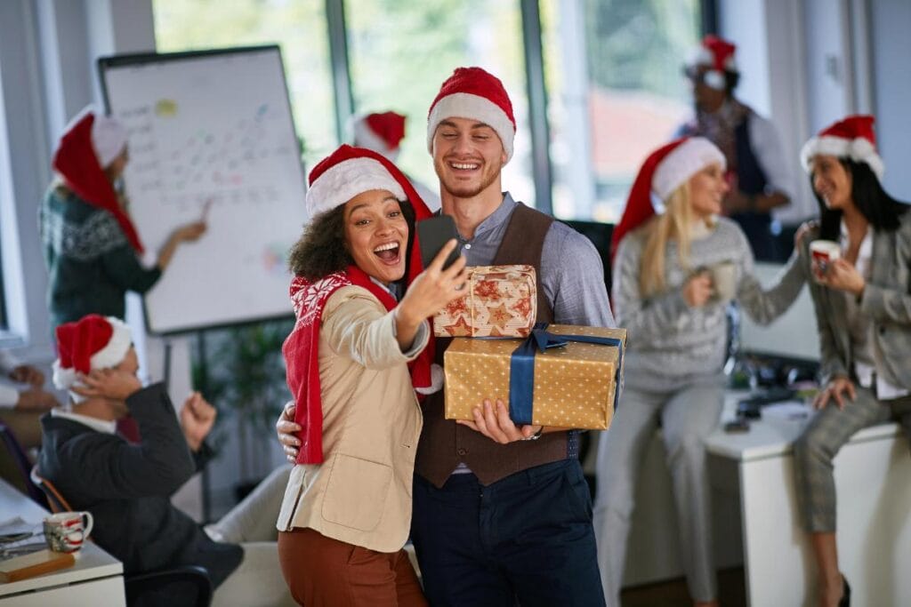 Coworkers in Santa hats enjoying a lively office Christmas party, with two people taking a selfie while holding gifts, and others chatting in the background.