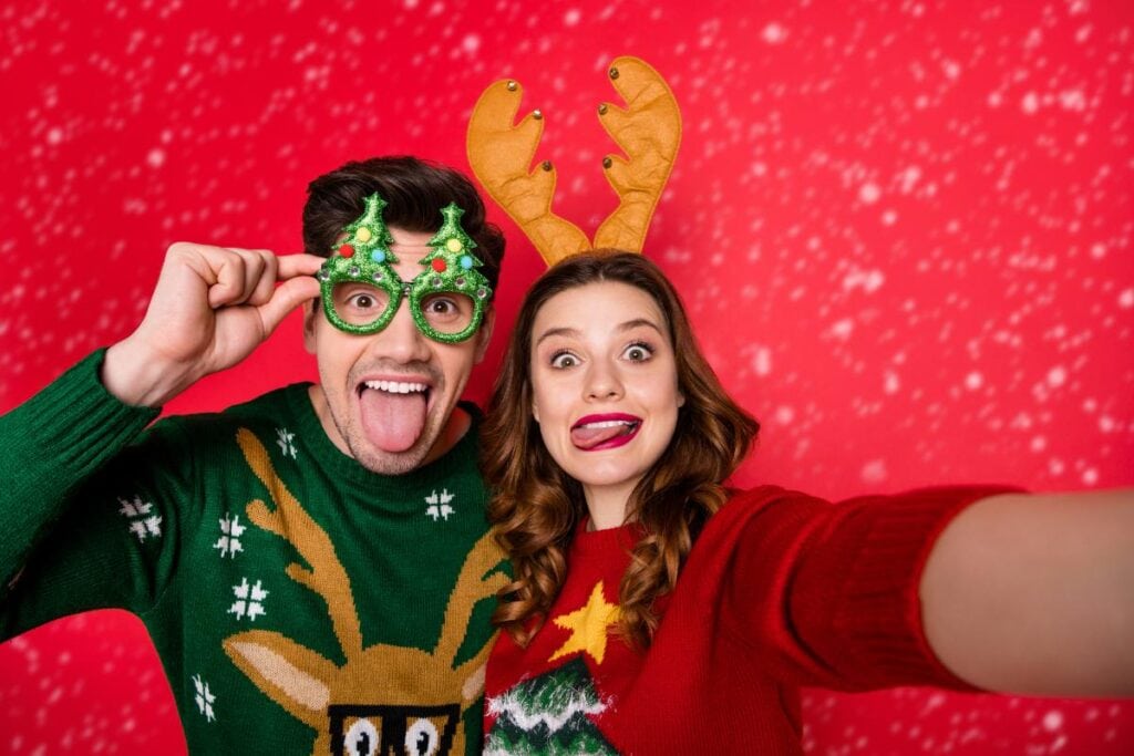 Smiling couple in Christmas sweaters making silly faces while taking a selfie in front of a red holiday backdrop with snowflake patterns.