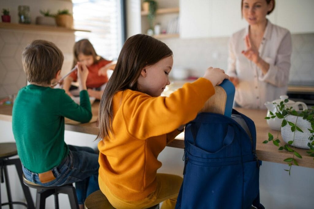 Three children at a kitchen counter during a busy morning routine, with a mother in the background giving instructions.