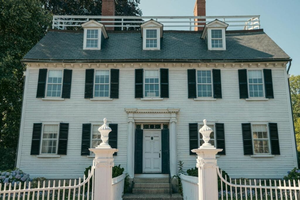 A stately white Colonial house with black shutters, a classic symmetrical facade, and a decorative white gate leading to the front steps.