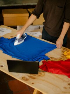 A person ironing a blue t-shirt on a wooden table, surrounded by colorful shirts, boxes, and shipping materials.