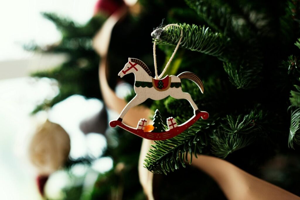 Wooden rocking horse ornament decorated with holiday accents, hanging on a Christmas tree branch with ribbon in the background.