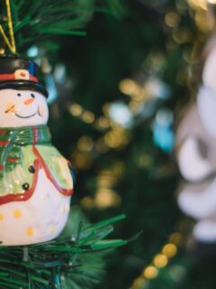 Close-up of a cheerful ceramic snowman ornament with a green coat and red scarf hanging on a decorated Christmas tree.