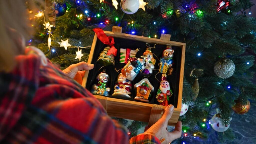 Person holding a wooden box filled with colorful, collectible Christmas ornaments in front of a lit Christmas tree.