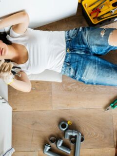 a woman in white shirt and blue jeans underneath the kitchen sink looking at the pipes and plumbing