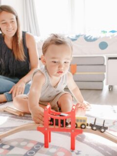 woman who could be an au pair sitting on floor watching a toddler boy play with a train set