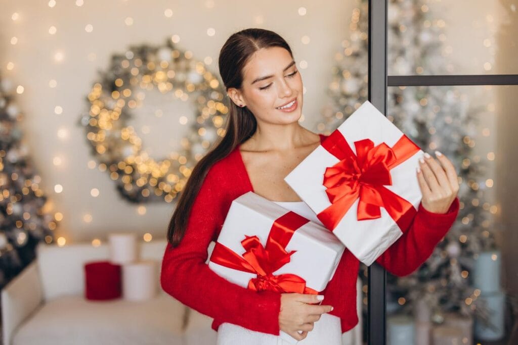 Woman dressed in red holding two white presents with red bows, standing in a softly lit room decorated with Christmas wreaths and lights.