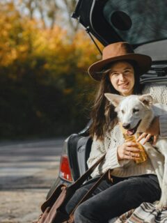 Smiling woman in a cozy sweater sits in the open trunk of a car, holding her happy white dog and a travel cup during a fall road trip break.