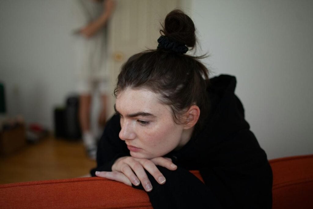 A person leaning on the back of a couch, looking down with a serious and contemplative expression, suggesting emotional strain or deep thought.
