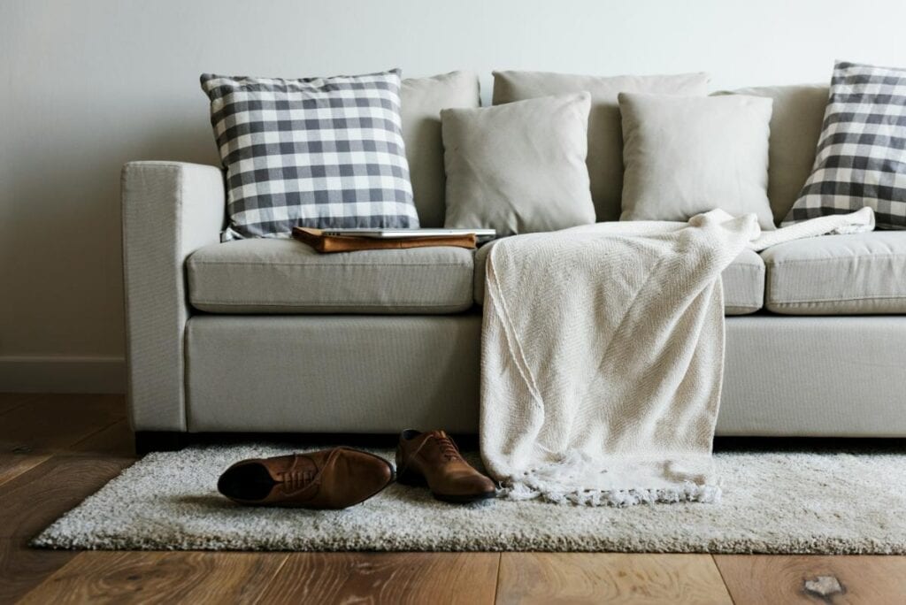 A neutral-toned sofa with layered throw pillows and a soft blanket sits on a textured rug, with brown leather shoes placed neatly in front.