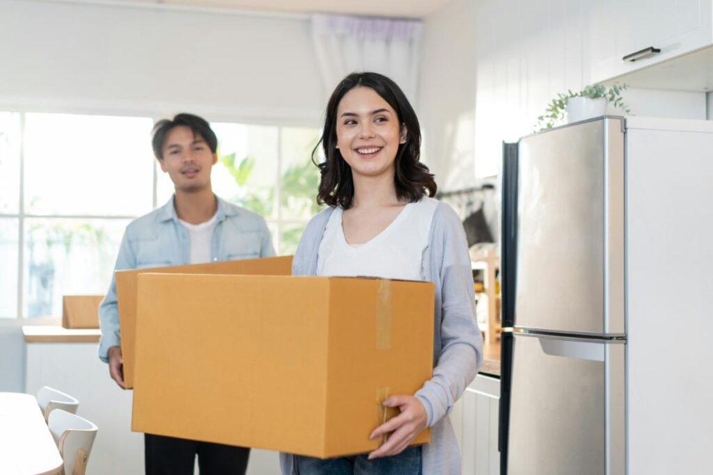 Smiling couple carrying cardboard boxes into a bright kitchen, moving into their new home.