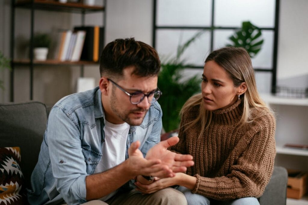 A distressed couple sits on a couch during a serious conversation, with the woman listening attentively as the man expresses frustration.