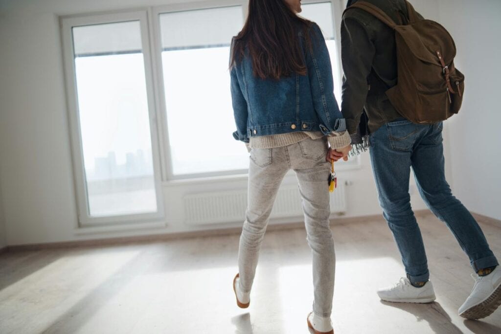 Couple holding hands and entering an empty apartment, holding keys and walking across a sunlit floor.