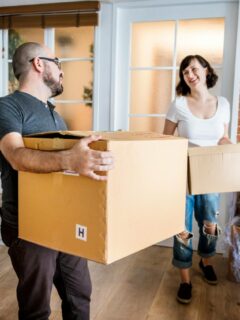 Man and woman carrying moving boxes into a cozy home with brick walls, smiling at each other as they unpack.