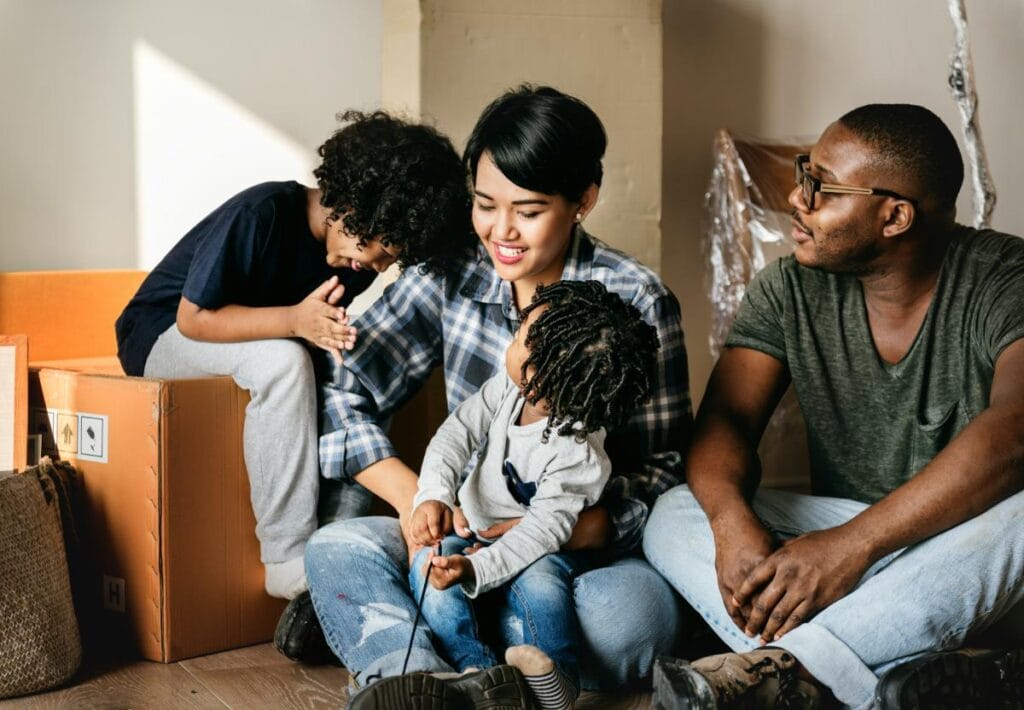 A smiling family sits on the floor surrounded by moving boxes, sharing a joyful moment together in their new home.