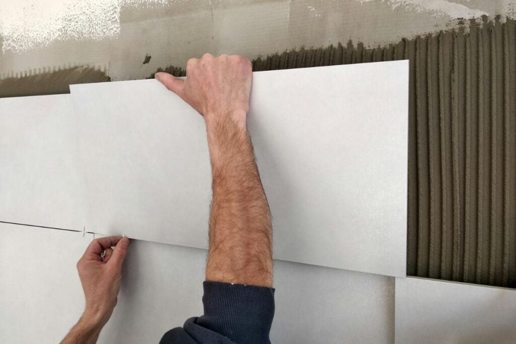 Hands installing large wall tiles with adhesive during a bathroom remodeling project, showing careful tile placement.