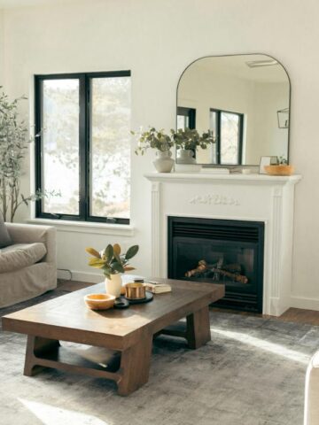 A light-filled living room showcases a cozy, well-styled space with soft armchairs, a wooden coffee table, minimalist accessories, and a large mirror above the fireplace.