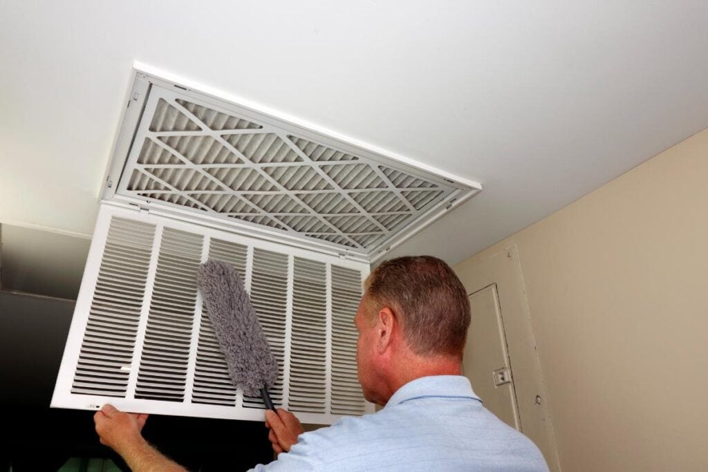 A technician cleaning an HVAC return vent with a duster while inspecting an air filter mounted on the ceiling.