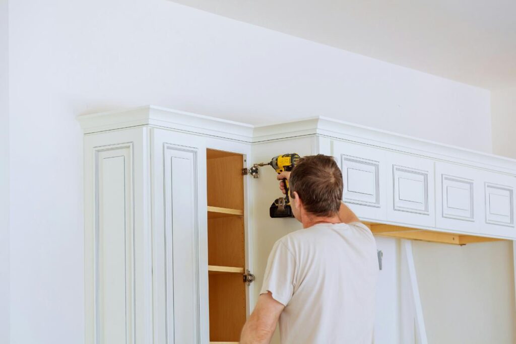 A man uses a power drill to install cabinet doors on upper kitchen cabinetry, highlighting the hands-on work of a kitchen remodel in progress.