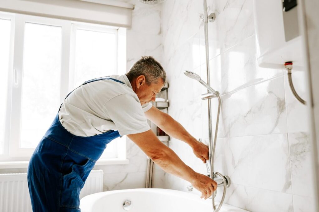 A man in blue overalls installs a handheld showerhead in a white tiled bathroom, focusing on practical plumbing updates during a remodel.