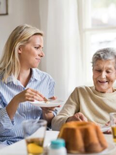 A smiling elderly woman enjoys a meal with her adult daughter at a sunlit dining table, showing warmth and shared care in daily life.