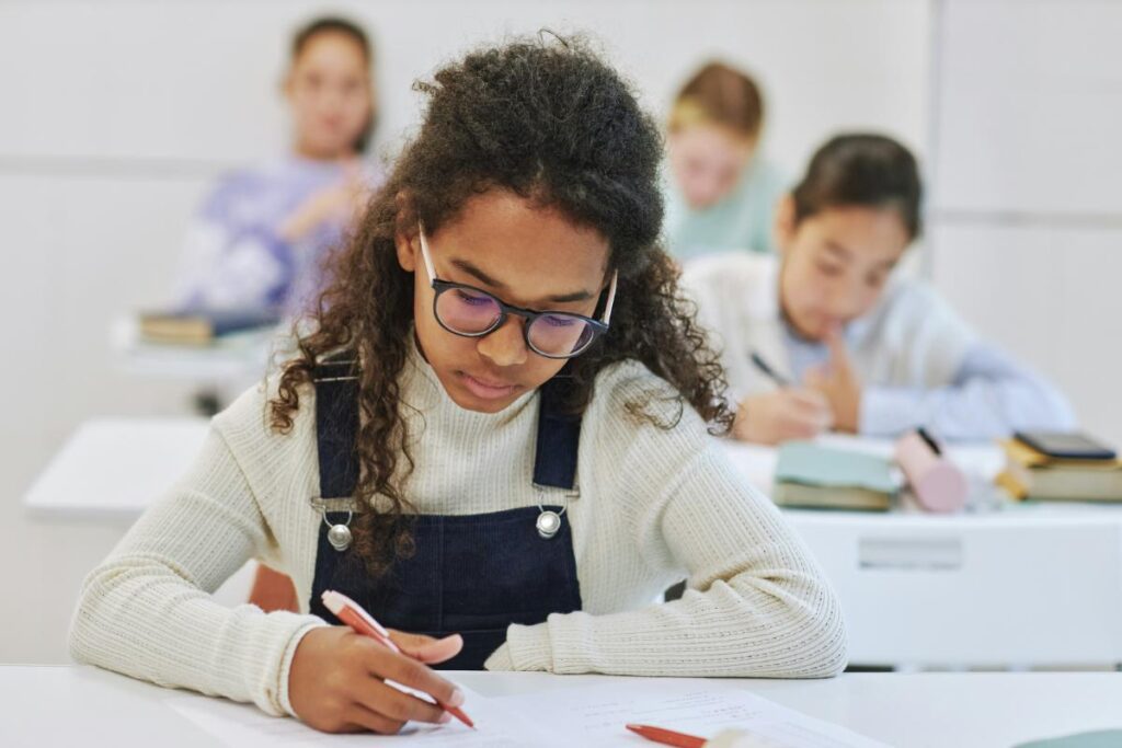 A teenage girl wearing glasses and a white sweater focuses intently on her schoolwork in a classroom setting with classmates in the background.