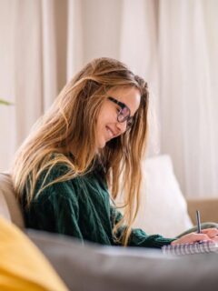 A smiling teenage girl with glasses sits on a sofa at home, writing in a journal with a pen, surrounded by cozy decor and natural light.