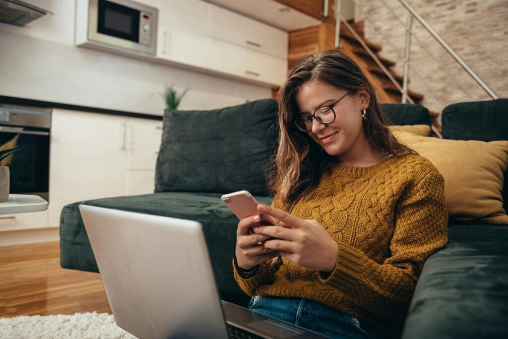 A teenage girl wearing glasses and a mustard sweater sits on the floor with a laptop in front of her, smiling as she looks at her phone.