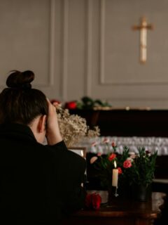 A grieving woman sits with her head in her hands near a closed casket adorned with flowers and candles in a quiet funeral setting.