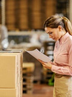 A woman in a warehouse checks inventory on a clipboard while referencing a laptop placed on stacked cardboard boxes, representing 3PL logistics management.