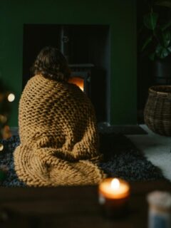A person wrapped in a thick blanket sitting near a lit fireplace, enjoying warmth and comfort in a cozy living room.